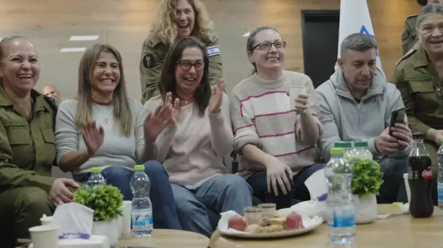 Three women in street clothes and three in military uniforms sit together, cheering and laughing