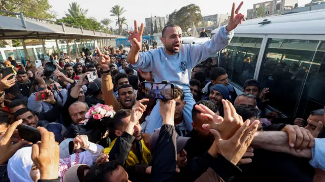 A released Palestinian prisoner is being carried on someone's shoulders above a crowd next to a white bus. He is doing a peace sign with both of his hands and he is wearing grey prison clothes.