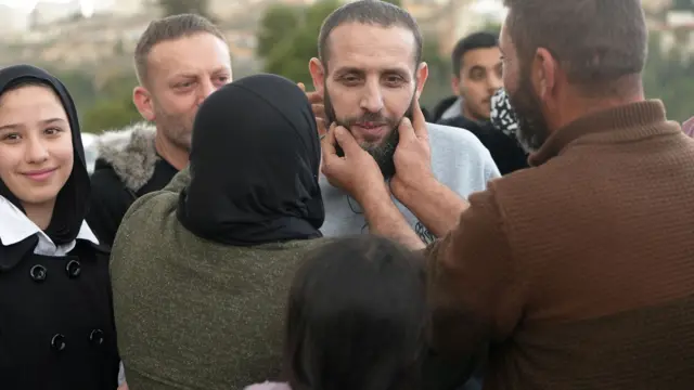 A woman wearing a black hijab and a man wearing a brown jumper put their hands on a released Palestinian prisoner's face. The released Palestinian prisoner is smiling and is wearing grey prison clothes.