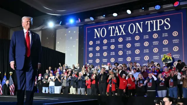 Trump smiles on stage, with a crowd behind him and "no tax on tips" written in large letters behind where he was speaking