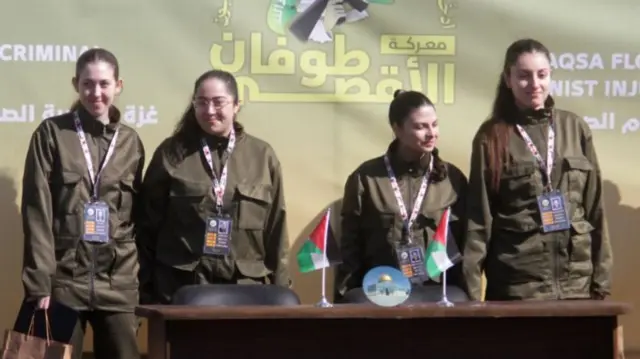 Four freed hostages in green military uniforms stand next to each other on a stage behind a wooden desk with two small Palestinian flags on top