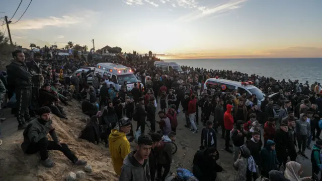 A crowd on a road with the sea in the background. Some vehicles are trying to get through