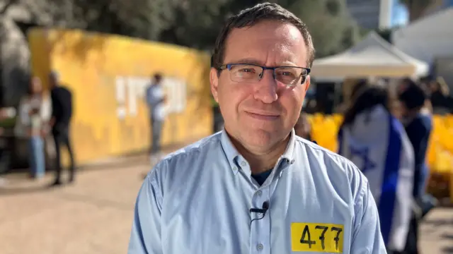 Professor Levine stands in Hostages Square in Tel Aviv. He is wearing wire-frame glasses and a blue shirt with a yellow sticker.