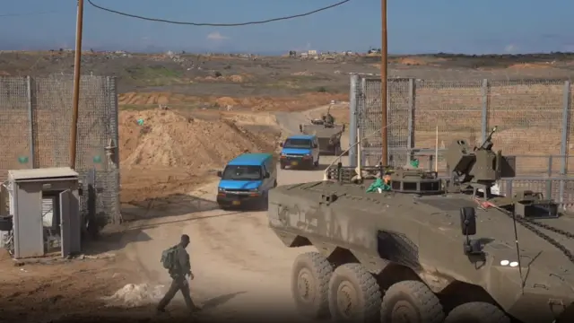 Two armoured vehicles escort two blue vans from Gaza back into Israel. A lone soldier is seen walking on the left side of the frame