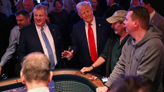 Donald Trump, in a blue suit and red tie, is surrounded by a crowd at a craps table in a Las Vegas casino