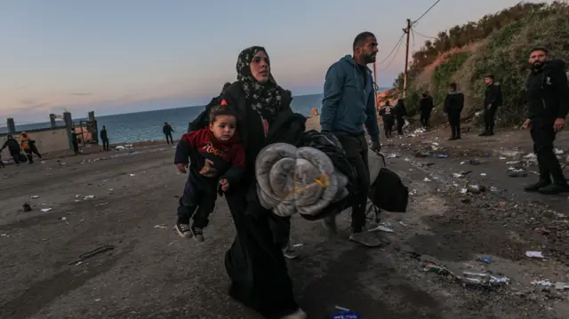 A woman holds a toddler while carrying rolled up blankets. A man walks next to her carrying bags. There is litter on the floor, and the sea is in the background.