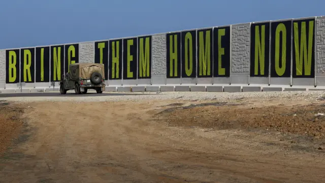 An Israeli military truck drives past a banner that reads 'bring them home now'