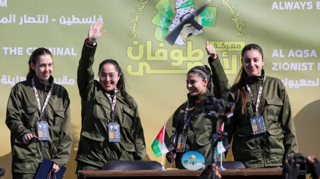 Four liberated hostages on stage wave and smile. They're in green military uniforms and are holding a black folder. There's a wooden table in front of them with two small Palestinian flags