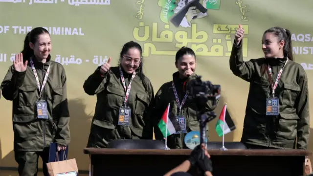 Four freed female soldiers in green military uniforms hold their thumbs up as they smile to the crowd