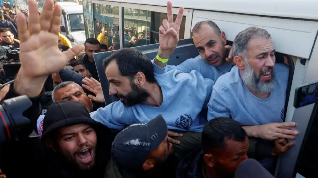 Three released Palestinian prisoners in a white bus lean out of the window, where there is a crowd. One of them is doing a peace sign with his right hand, and all three of them are wearing grey prison clothes.