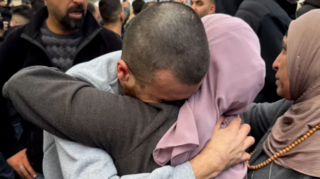 Release Palestinian prisoner in grey prison clothes has an emotional hug with a woman wearing a pink hijab. They are both in a crowd.