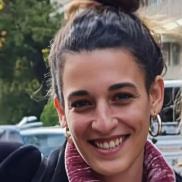 Arbel Yehud, smiling at camera, wearing earrings and hair tied back, photographed from shoulders up