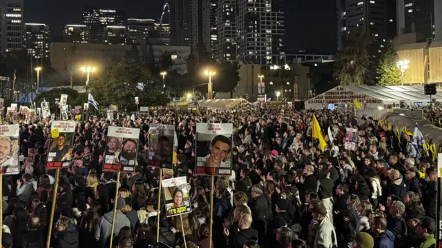 A crowd of people in a large open area in the city of Tel Aviv at night. Some are holding up placards with the faces of some of the Israeli hostages.