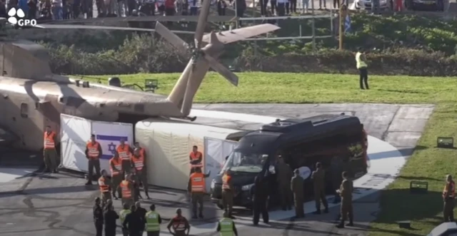 Tail end of helicopter with people in high vis vests holding a shield around the aircraft's exit with a black van