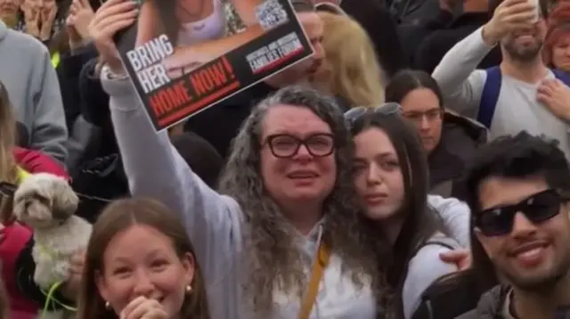 Family and friends of the hostages in tears and smiling as they celebrate. One woman with long grey hair and black rimmed glasses holds up a poster with a photo of one of the hostages