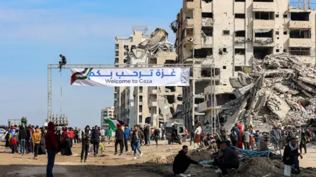 People gather by a banner welcoming people near the rubble of a collapsed building along Gaza's coastal al-Rashid Street for people to cross from the Israeli-blocked Netzarim corridor from the southern Gaza Strip into Gaza City on January 26, 2025