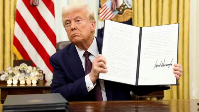 Donald Trump holding up a signed executive order at his desk in the Oval Office