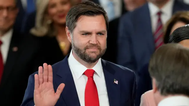 JD Vance holds up his right hand as he is sworn in as sworn in as vice-president. He has blue eyes, brown hair and a closely-trimmed brown beard. He is wearing a navy blazer, white shirt, and red tie.