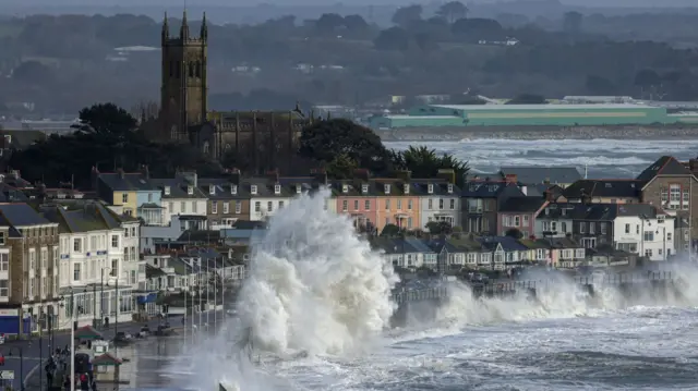 Waves seen crashing up onto the pathway and roads along Penzance with rows of buildings and a church at the top of a hill. Mainland can be seen further afield.