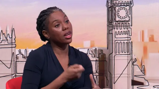 Kemi Badenoch in black shirt sits down on red chair, her right hand slightly extended in front of her