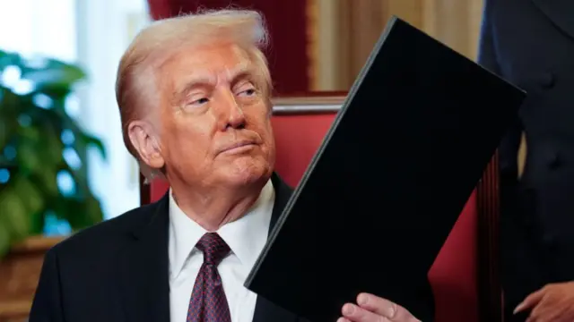 A seated Trump hands over a black folder containing an executive order to an aide who is standing outside of the frame