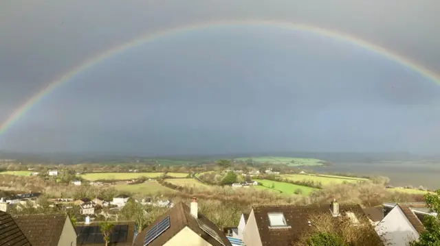 Grey skies with a whole rainbow above fields and houses.