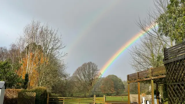 A double rainbow seen with trees in front, taken from a garden with fencing in front.