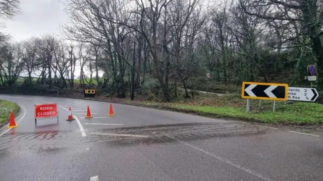 A road closed with orange cones and a road closed sign stopping traffic. Water can be seen in a stream across the road.