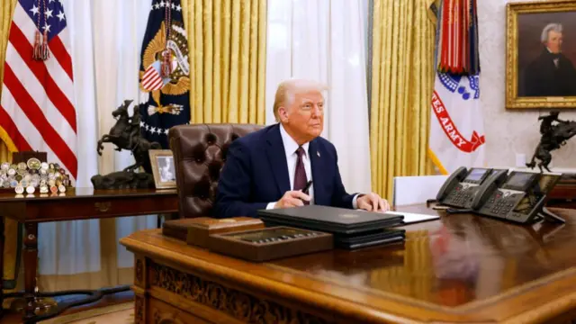 Trump, wearing a blue suit with purple tie, sits at the Resolute Desk in the Oval Office