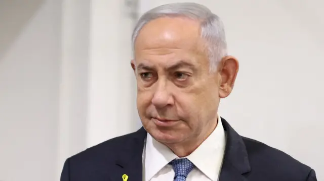 Benjamin Netanyahu looks on. He has thin, short white hair. He is wearing a black blazer, white shirt, and a light blue tie.