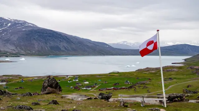 Greenland's flag (rectangle evenly divided in two, with white top and red bottom. A circle in the middle, split in two, with red top and white bottom) flies in Igaliku settlement. Large expanse of grass on a hill, with a lake and a mountain range in the background