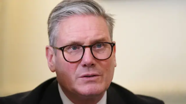 Keir Starmer close up, he's sitting down and wearing a black suit and white shirt with black-rimmed glasses, a white wall in the background