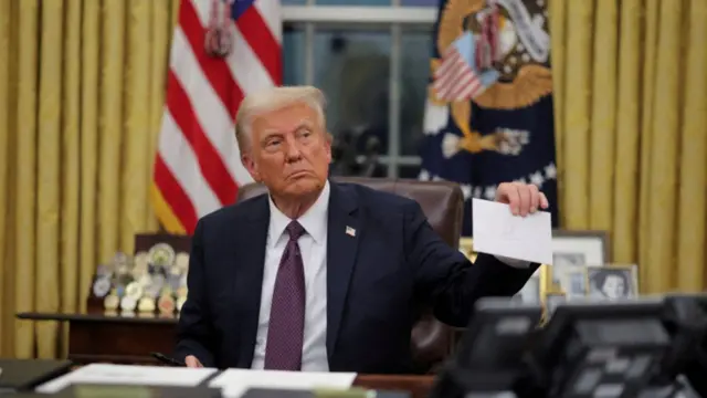 Joe Biden holds up a white envelope bearing the signature of Joe Biden while he sits down at his desk in the White House