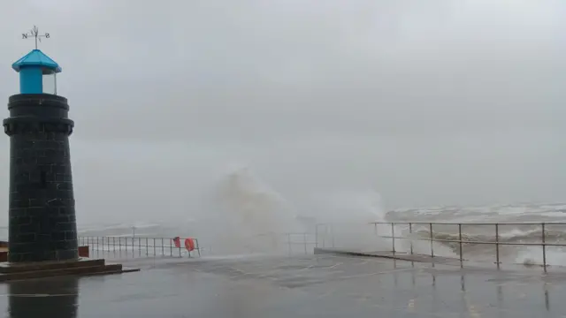 Waves crashing onto the path with a small lighthouse on the left on land.