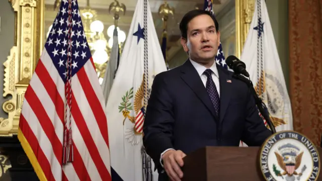 Marco Rubio stands behind a stand and microphone. An American flag and other flags stand behind him.