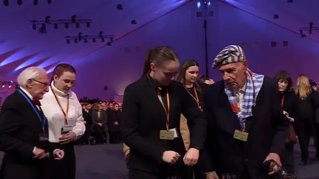 Crowds of people are gathered wearing lanyards. One wears a scarf and hat that has blue and white stripes.