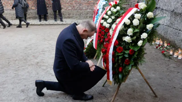 Polish President Andrzej Duda, wearing all black, kneels before two wreaths placed in front of a wall. Candles have also been placed in front of the wall.