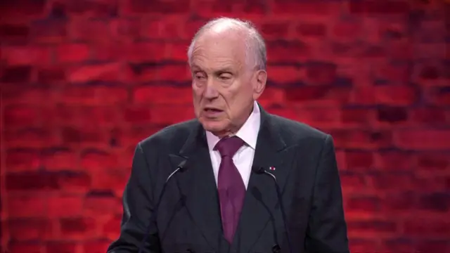 Ronald Lauder wears a black suit and purple tie speaking behind a podium with microphones.