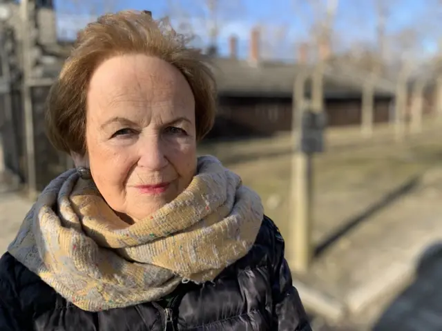 Mala Tribich looking at camera, wearing black jacket and light-coloured scarf with Auschwitz camp blurred in background