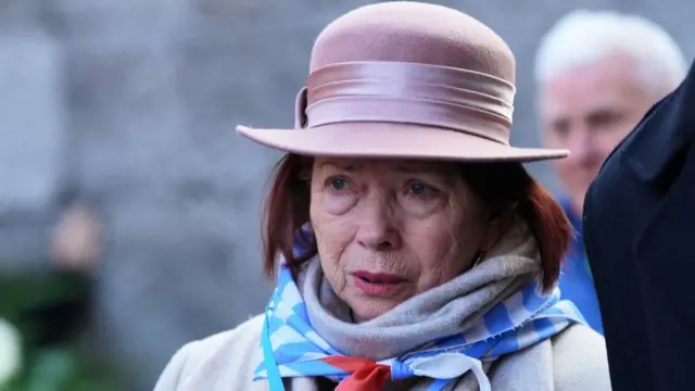 A woman wearing a pink hat looks upset as she watches the commemorations. She is wearing a blue and white striped scarf