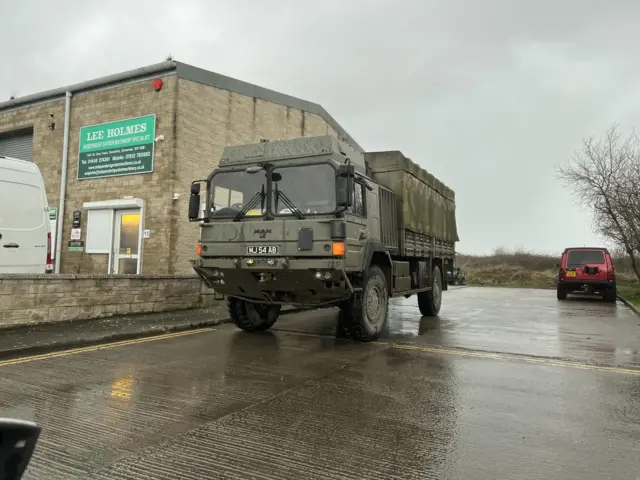 A large khaki green military truck parked on a quiet road on an industrial estate. The sky is grey and quite dark with clouds, and there is rainwater on the road.