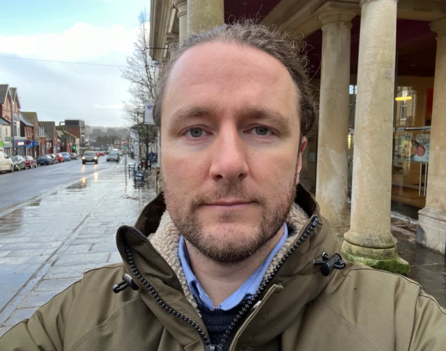 The image shows reporter Ross Pollard looking into the camera. The Guildhall in Chard is in the background.