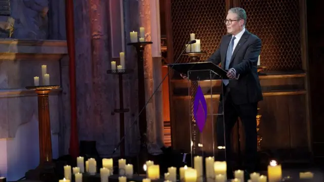 Starmer, wearing a suit, stands at a lectern decorated with a purple flame, speaking