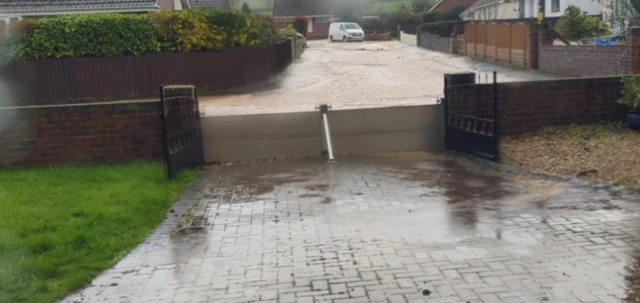 The picture shows a flood barrier across a drive with rain water building on the road