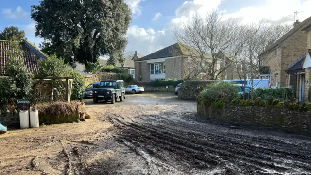 A muddy driveway in front of Burnworthy House. Multiple tyre tracks can be seen in the muddy road