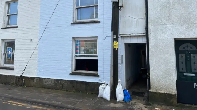 The view of a residential street where a house door hangs open with a few sandbags outside