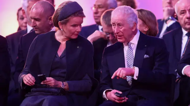 King Charles and Queen Mathilde speak while sitting among others at the event