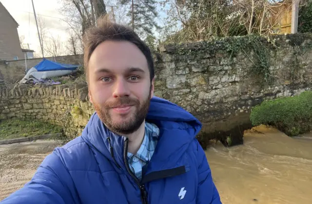 BBC reporter Dan Ayers looking at the camera, wearing a royal blue waterproof jacket with floodwater in the background