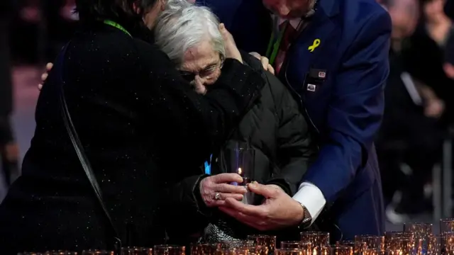 An old woman leans into the arms of another as she emotionally places a candle down amongst others