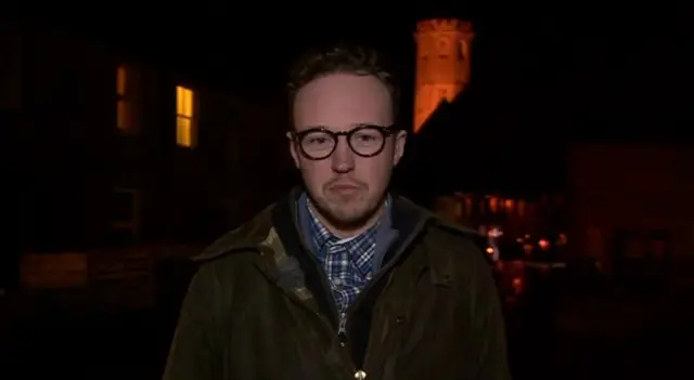 MP Adam Dance, looking directly at the camera while being interviewed live by the BBC. He is standing on a dark street with a church lit up behind him, and wears a blue checked shirt, a fleece and a dark work jacket. He also has round black glasses on.